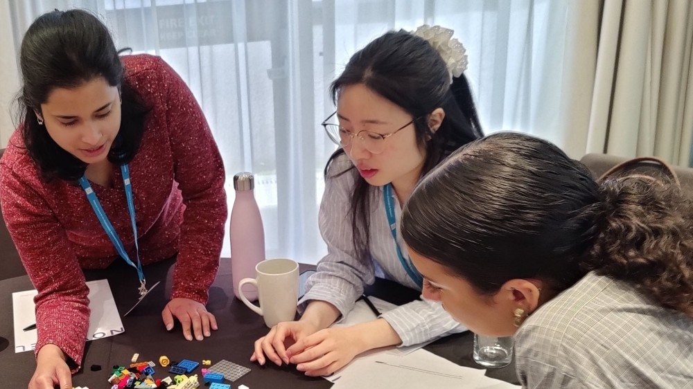 Three students around a table in discussion over some lego bricks.
